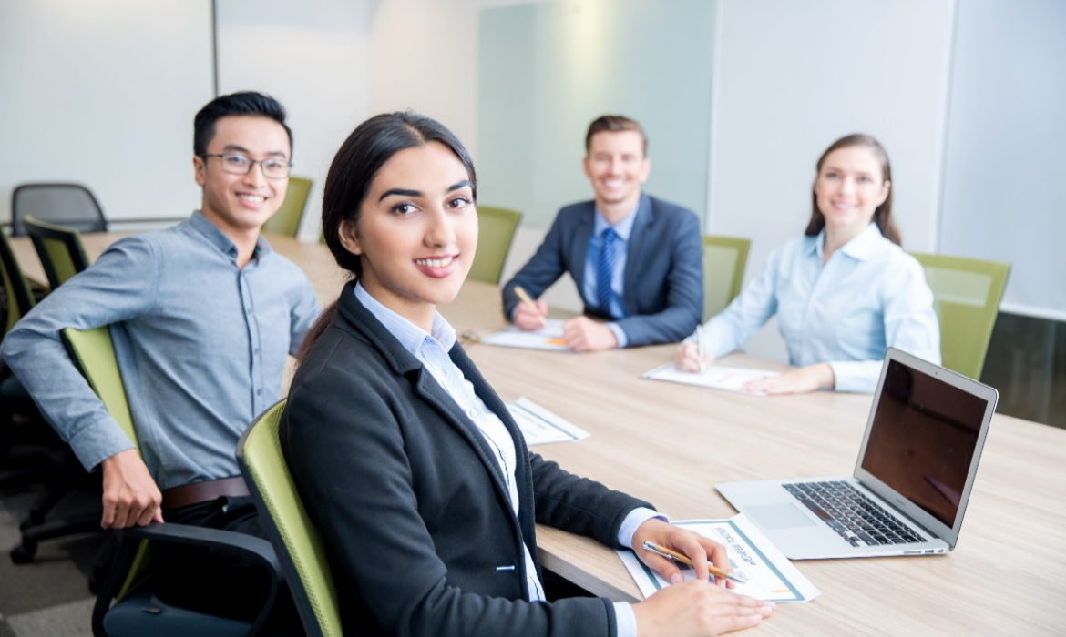 Four smiling middle-aged multi-ethnic business people turning to camera, working and discussing ideas while sitting at big table in conference room. Side view.